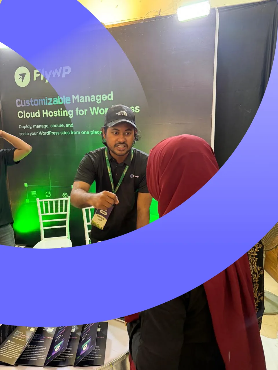 A FlyWP team member in a black FlyWP polo speaking with a visitor in front of the booth banner